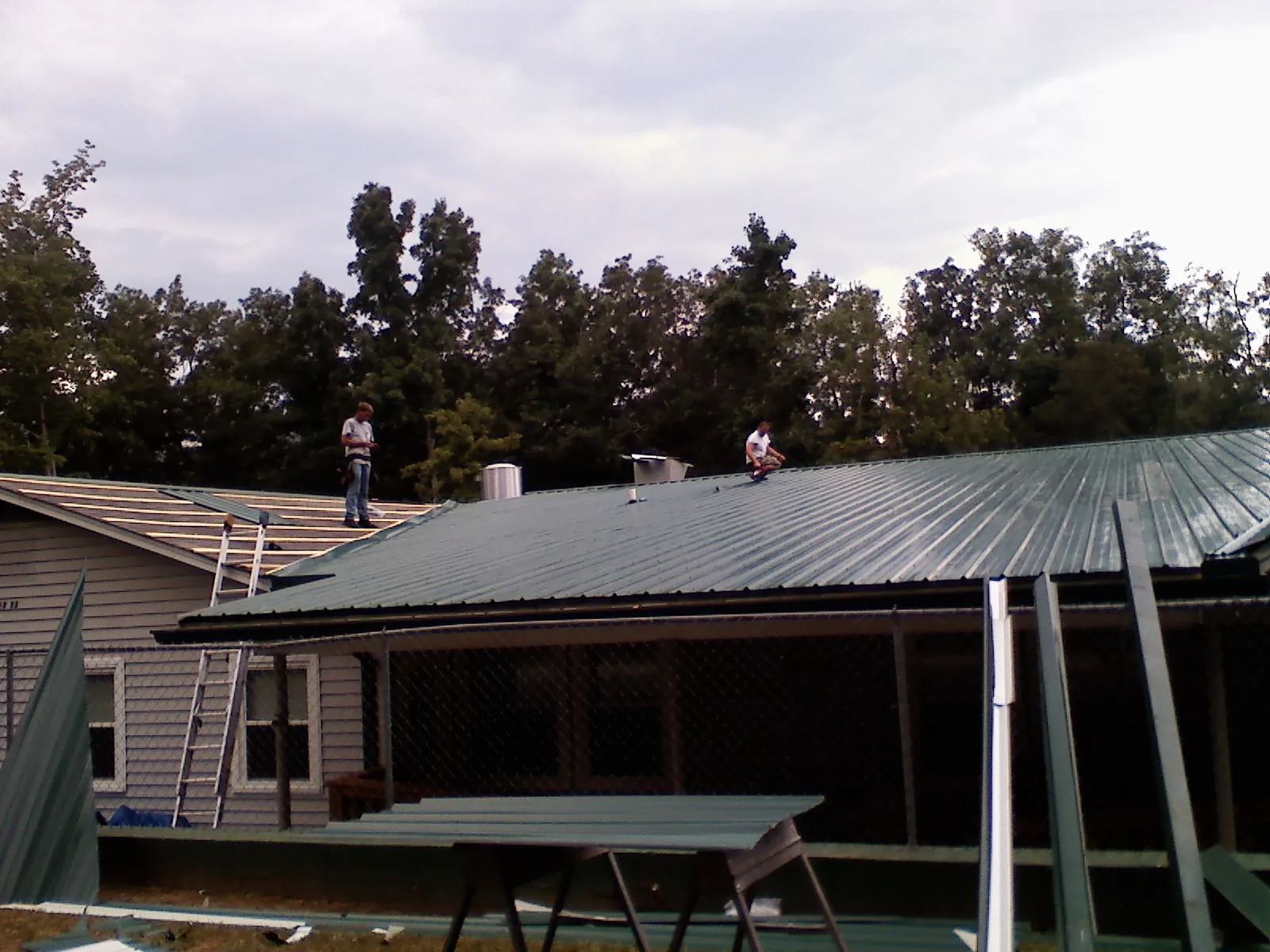 Two roofers installing green metal panels above a covered porch in Maple City